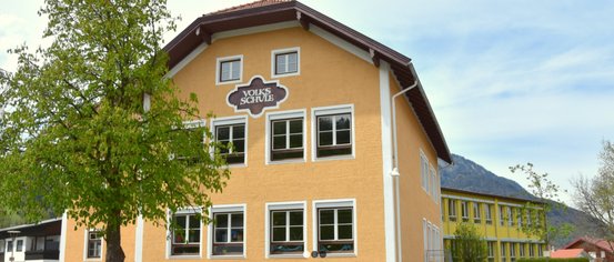 Ein Bild eines gelben Gebäudes mit mehreren Fenstern. Der Name 'Volksschule' ist über dem Eingang angebracht.