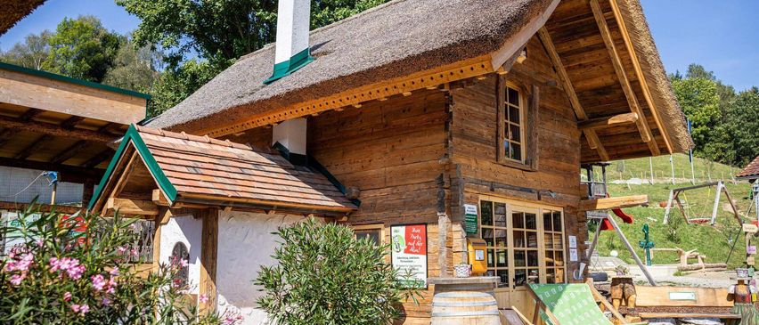 Ein Holzhaus mit Strohdach, Schornstein und Glastür. Ein Topf und ein Fass stehen davor.