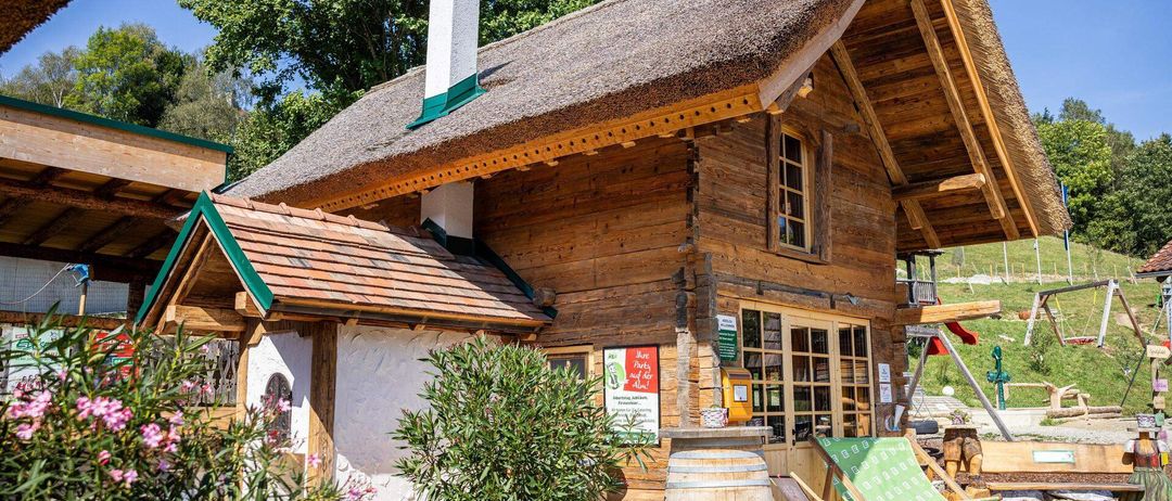 Ein Holzhaus mit Strohdach, Schornstein und Glastür. Ein Topf und ein Fass stehen davor.