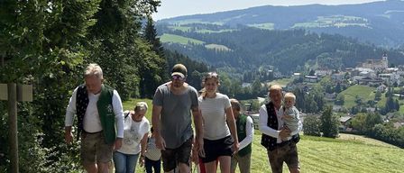 Eine Gruppe von Menschen läuft auf einem Kiesweg, umgeben von üppiger Vegetation und Bergen. Der Mann in der Mitte trägt einen Hut und Sonnenbrille. Ein Mann auf der rechten Seite hält ein Baby.