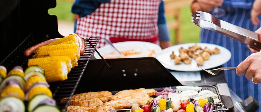 Ein Grill im Freien mit verschiedenen Speisen, darunter gegrilltes Hähnchen, Gemüse, Maiskolben und Würstchen. Eine Person in einer rot-weiß karierten Schürze steht hinter dem Grill.
