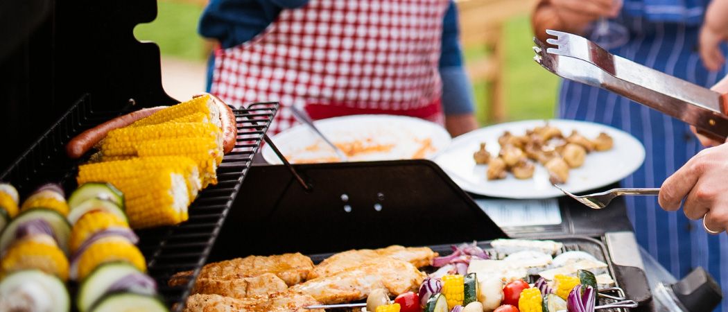 Ein Grill im Freien mit verschiedenen Speisen, darunter gegrilltes Hähnchen, Gemüse, Maiskolben und Würstchen. Eine Person in einer rot-weiß karierten Schürze steht hinter dem Grill.