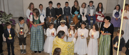 Ein Priester steht vor einer Gruppe von Kindern in traditioneller Kleidung und hält brennende Kerzen. Die Kinder in weißen Kleidern sind um den Priester versammelt. Ein Tisch mit Kerzen steht in der Mitte.