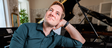 Ein Mann mit blauen Augen sitzt neben einem Klavier, trägt ein grünes Hemd und hält sich mit der Hand ans Kinn.