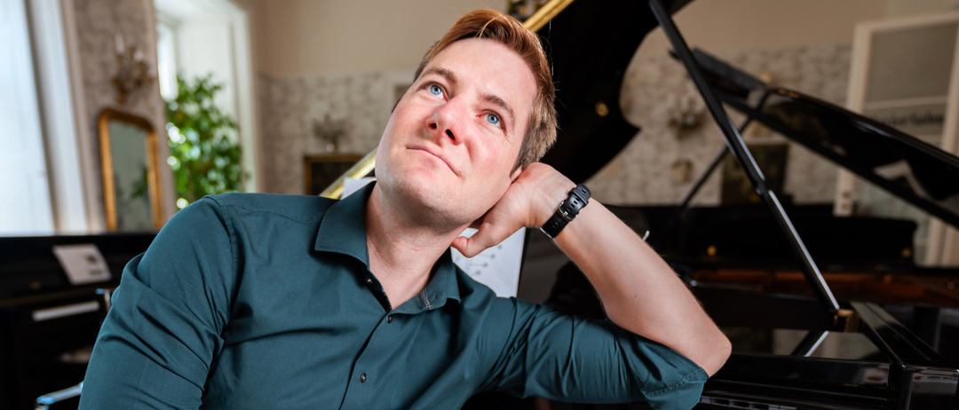 Ein Mann mit blauen Augen sitzt neben einem Klavier, trägt ein grünes Hemd und hält sich mit der Hand ans Kinn.