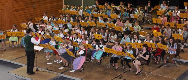 Eine große Gruppe von Kindern und Erwachsenen spielt auf der Bühne mit verschiedenen Instrumenten, darunter Flöten und Posaunen, unter der Leitung eines Dirigenten. Das Publikum sitzt im Hintergrund.