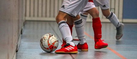 Soccer players in red and white uniforms with red shoes play indoors, with a soccer ball at their feet.