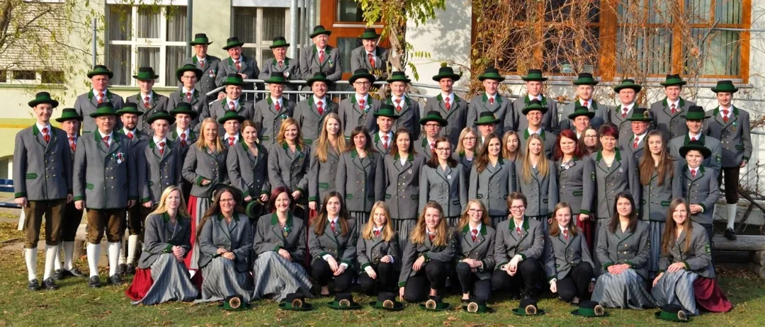 Eine Gruppe von Personen in traditioneller Kleidung mit grünen Hüten, die vor einem Gebäude mit herbstlicher Dekoration stehen und sitzen.