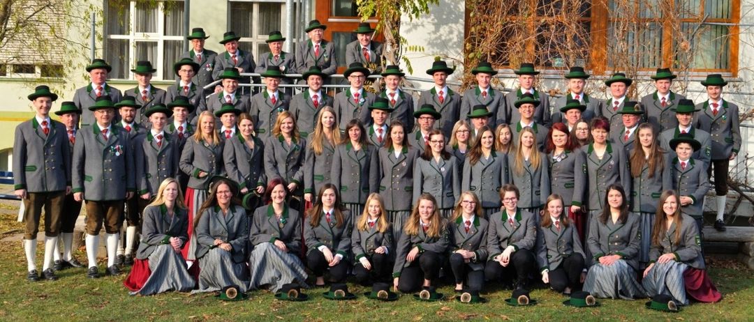 Eine große Gruppe von Menschen in traditionellen Uniformen posiert für ein Foto vor einem Gebäude. Sie sitzen und stehen auf dem Rasen, mit Bäumen und Pflanzen im Hintergrund.
