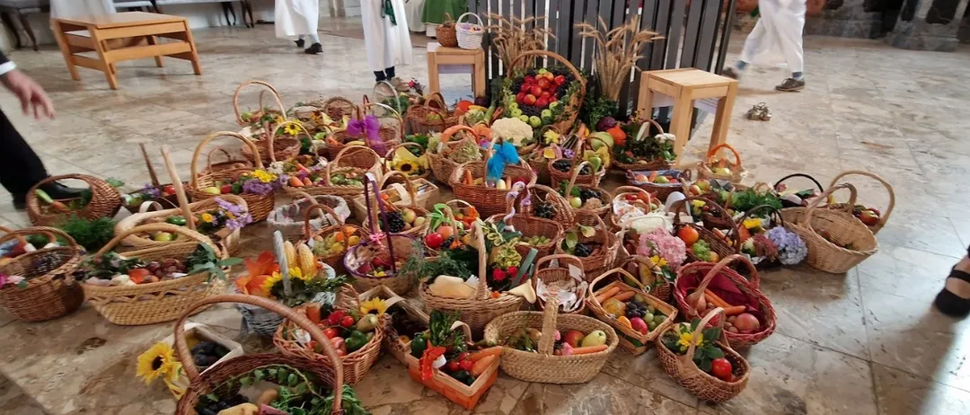 Mehrere Menschen in weißer Kleidung stehen hinter einem mit Blumen und Früchten geschmückten hölzernen Altar. Der Altar ist mit einer Kerze und einem Buch geschmückt. Verschiedene Körbe mit Obst und Gemüse sind auf dem gefliesten Boden ausgestellt.