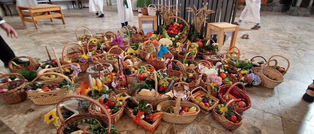 Mehrere Menschen in weißer Kleidung stehen hinter einem mit Blumen und Früchten geschmückten hölzernen Altar. Der Altar ist mit einer Kerze und einem Buch geschmückt. Verschiedene Körbe mit Obst und Gemüse sind auf dem gefliesten Boden ausgestellt.