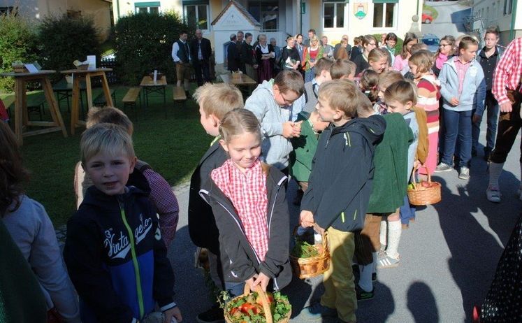 Eine Gruppe von Kindern und Erwachsenen versammelt sich vor einem Gebäude. Einige Kinder halten Körbe mit Erdbeeren. Die Erwachsenen stehen im Hintergrund.
