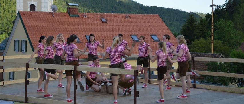 Eine Gruppe von Frauen in traditionellen Kostümen tanzt auf einer Holzplattform, mit einem rotgedeckten Gebäude und Bergen im Hintergrund.