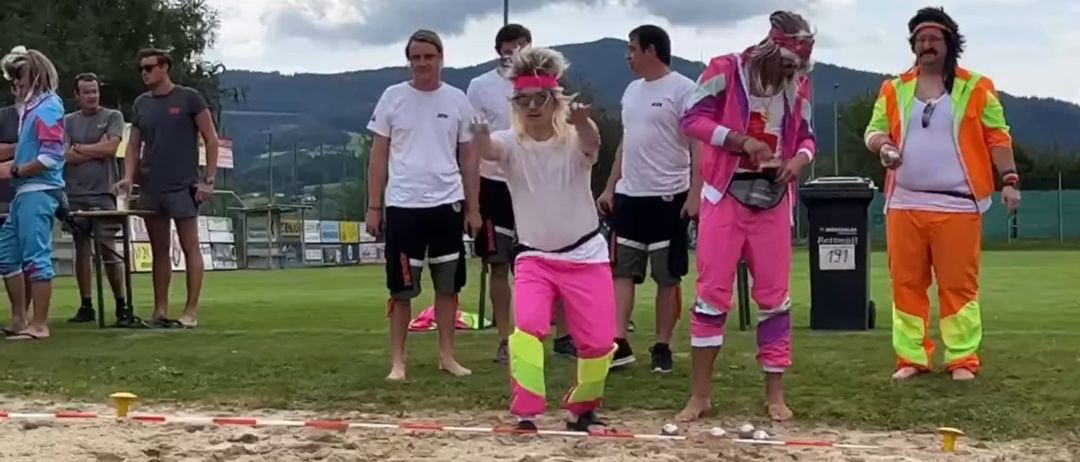 Eine Gruppe von Menschen in einem Park, eine Person in Pink und Weiß steht auf einem Sandweg, während andere zusehen.