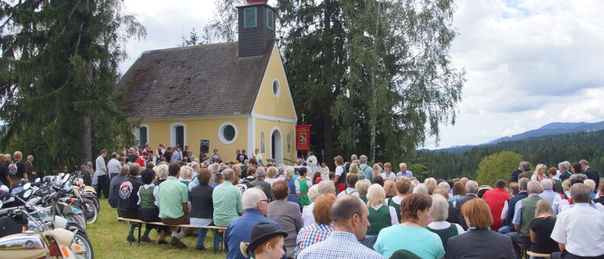 Eine Menschenmenge, einschließlich Kinder, sitzen auf Bänken vor einer gelben Kirche mit einem roten Kuppel. Sie scheinen einen Gottesdienst zu besuchen.