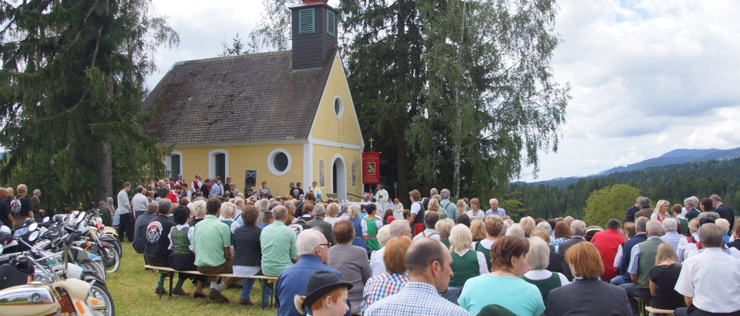 Eine Menschenmenge, einschließlich Kinder, sitzen auf Bänken vor einer gelben Kirche mit einem roten Kuppel. Sie scheinen einen Gottesdienst zu besuchen.