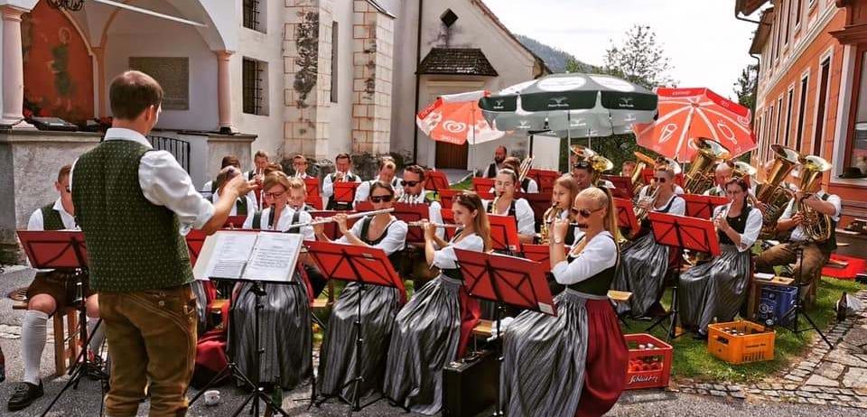 Ein traditionelles Orchester spielt vor einer Kirche, mit Musikern, die Flöten, Tubas und Posaunen unter roten Sonnenschirmen spielen. Der Dirigent leitet, und das Publikum sitzt auf dem Gras.