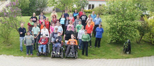 Eine Gruppe von Menschen ist in einem Park versammelt, einige in Rollstühlen. Bäume und Büsche umgeben sie, und ein orangefarbenes Gebäude ist im Hintergrund.