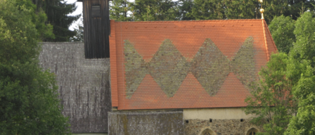 Eine kleine, alte Kirche mit einem grauen Dach und einem orangefarbenen Dach mit einem geometrischen Muster. Bäume und ein Zaun sind drumherum. Zwei Kühe grasen auf dem Rasen vor der Kirche.