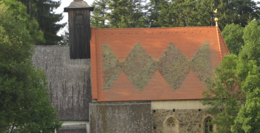 Eine kleine, alte Kirche mit einem grauen Dach und einem orangefarbenen Dach mit einem geometrischen Muster. Bäume und ein Zaun sind drumherum. Zwei Kühe grasen auf dem Rasen vor der Kirche.