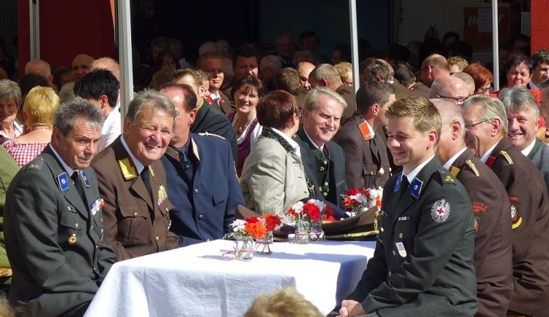 Eine Gruppe uniformierter Männer in Militärkleidung sitzt an einem Tisch mit Blumen in Vasen, umgeben von einer größeren Menschenmenge. Die Szene wirkt formell und festlich, mit vielen Menschen im Hintergrund.