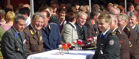 Eine Gruppe uniformierter Männer in Militärkleidung sitzt an einem Tisch mit Blumen in Vasen, umgeben von einer größeren Menschenmenge. Die Szene wirkt formell und festlich, mit vielen Menschen im Hintergrund.