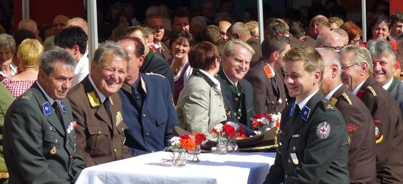 Eine Gruppe uniformierter Männer in Militärkleidung sitzt an einem Tisch mit Blumen in Vasen, umgeben von einer größeren Menschenmenge. Die Szene wirkt formell und festlich, mit vielen Menschen im Hintergrund.