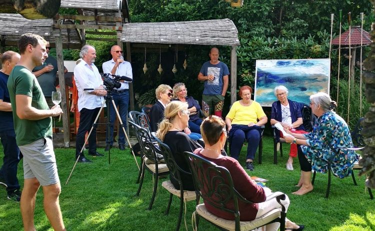 Eine Gruppe von Menschen sitzt in einem Garten mit Bäumen im Hintergrund. Eine Person hält ein Buch, eine andere steht mit einem Weinglas. Eine Kamera auf einem Stativ ist in der Nähe positioniert.
