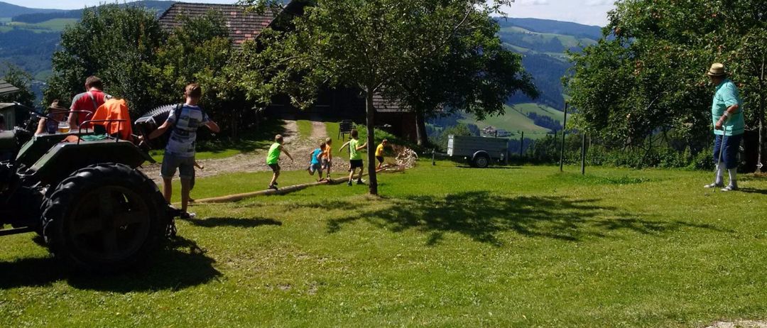 Eine Gruppe von Kindern rennt auf einem Grasfeld, während ein Mann ein Werkzeug hält. Ein Haus, ein Baum und ein Anhänger sind im Hintergrund.