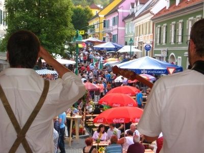 Zwei Männer mit Gitarren spielen bei einem lebhaften Straßenfest. Bunte Zelte und Schirme säumen die Straße, und viele Menschen genießen Essen und Getränke.