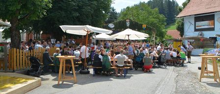 A large crowd is gathered outside an outdoor restaurant under white umbrellas, with people sitting on benches and tables. Trees and a fence are in the background, along with a mountain and a building.