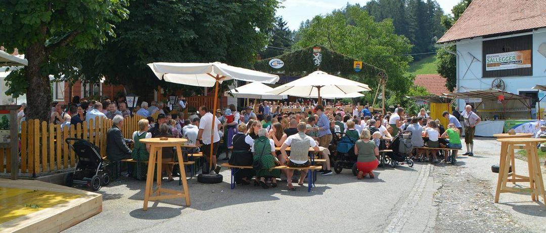 Eine große Menschenmenge versammelt sich unter weißen Sonnenschirmen vor einem Restaurant im Freien, wobei die Leute auf Bänken und Tischen sitzen. Im Hintergrund befinden sich Bäume und ein Zaun, sowie ein Berg und ein Gebäude.