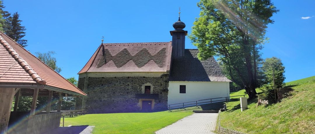 Eine alte Steinkirche mit Turm und Spitzdach, unter blauem Himmel, mit einem Weg, der zum Eingang führt.
