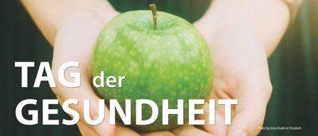Eine Hand hält einen grünen Apfel, mit dem Text 'der undheit' darunter in weiß geschrieben. Der Apfel ist leicht glänzend und hat ein paar Sprenkel.