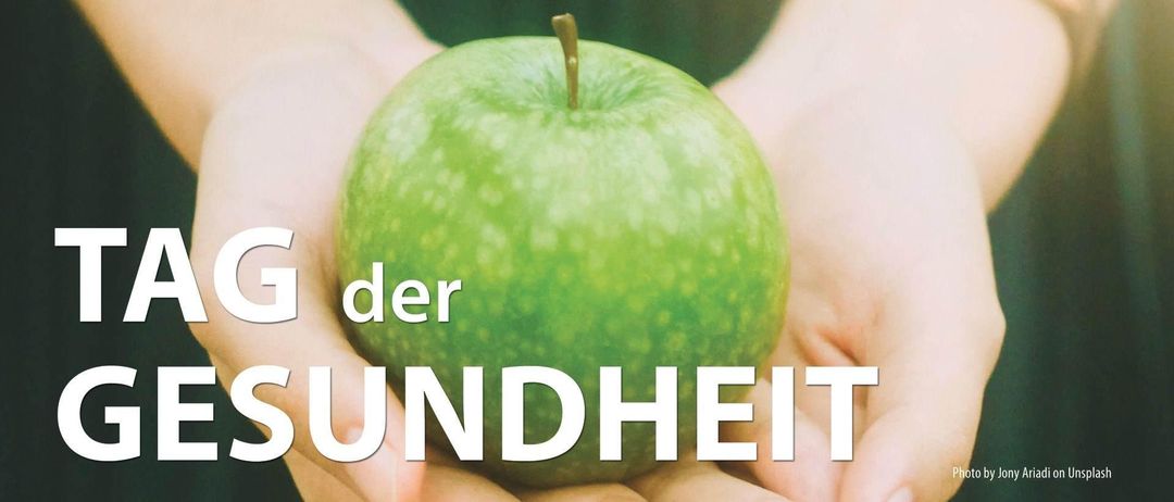 Eine Hand hält einen grünen Apfel, mit dem Text 'der undheit' darunter in weiß geschrieben. Der Apfel ist leicht glänzend und hat ein paar Sprenkel.