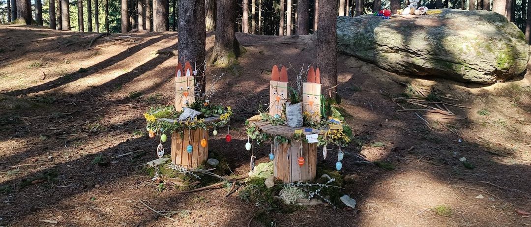 Zwei hölzerne Osterdekorationen stehen in einer Waldlichtung, umgeben von Bäumen und Laub. Jede hat einen Hasenkopf und einen kleinen Tisch, der mit Ostereiern und Blumen geschmückt ist.