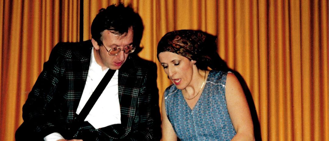 A man and woman are standing next to a large metal basin, the woman is washing something while the man holds a gun and looks at her.