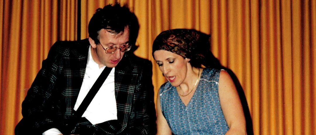 A man in a plaid suit and a woman in a blue dress are standing next to a large metal bucket, possibly performing a play.