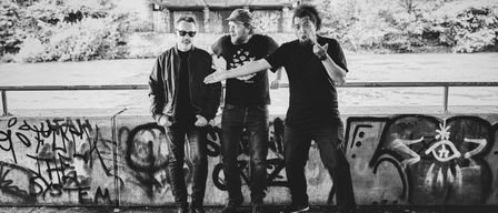 Drei Männer stehen auf einer Betonplattform, einer gestikuliert mit einer Hand, Graffiti an der Wand hinter ihnen und eine Brücke darüber.