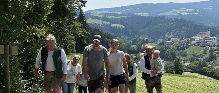 Eine Gruppe von Menschen geht auf einem Grasweg mit einer malerischen Aussicht auf Berge und ein Dorf. Sie sind lässig gekleidet, und einige tragen Kinder. Im Hintergrund zeigt die Landschaft Bäume, Häuser und einen klaren blauen Himmel.