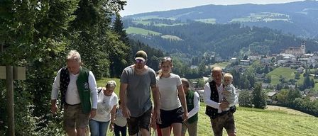 A group of hikers, including men and women, walk together on a grassy hillside, with mountains and a village in the background.