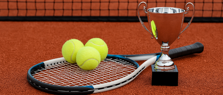 Ein Tennisschläger mit drei gelben Tennisbällen liegt auf einem roten Tennisplatz neben einem silbernen Pokal.