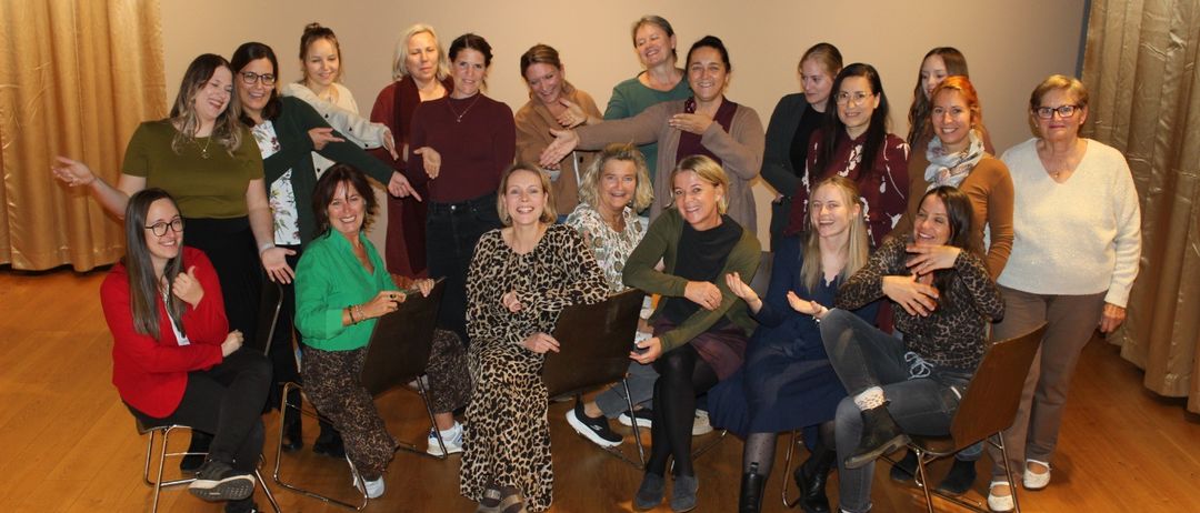 Eine Gruppe von Frauen in einem Raum mit Holzböden posiert für ein Foto. Sie sitzen und stehen, lächeln und lachen. Einige tragen Brillen, Halsketten, Armbänder und Sneakers.