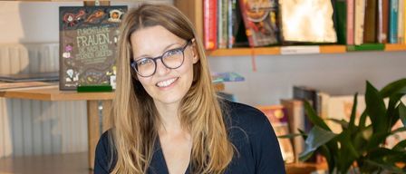 A woman sits on the floor, holding an open book, and smiling. She wears glasses and a dark blue outfit. Behind her, books are on a shelf and a potted plant is in a white pot.
