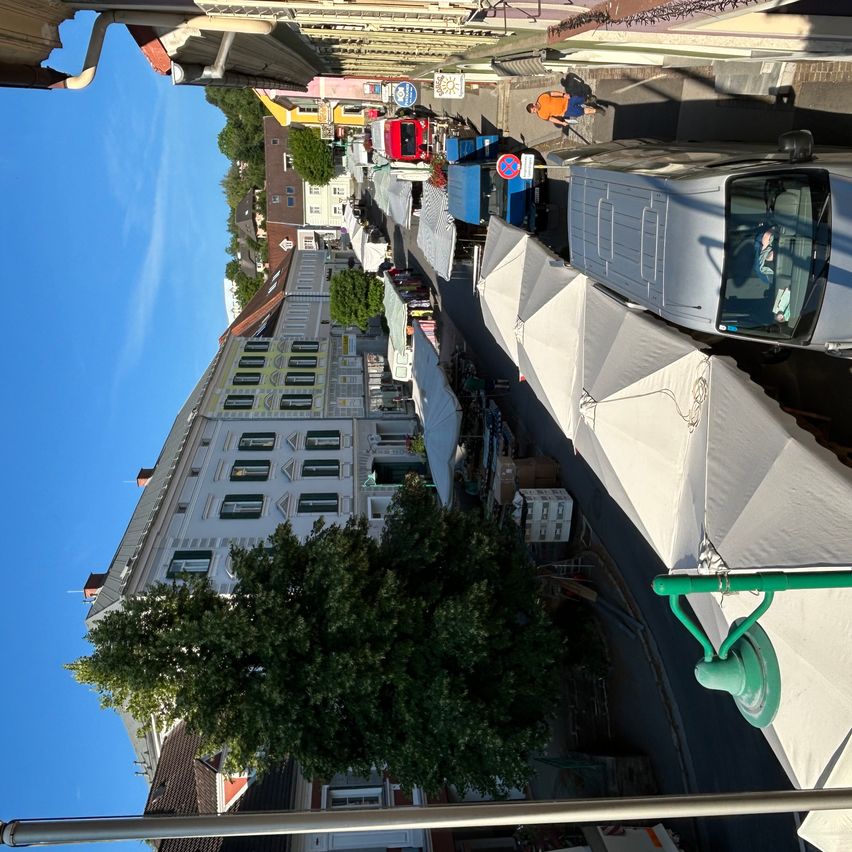 Luftaufnahme eines Straßenmarktes mit verschiedenen Ständen, einem vorbeigehenden Menschen und einem geparkten Van. Gebäude säumen die Straße, und ein Baum steht vor einem mehrstöckigen Gebäude.