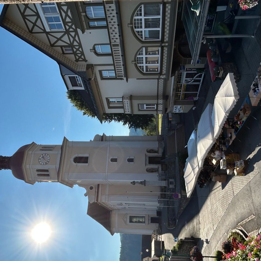 Eine Kirche mit Glockenturm steht unter einem strahlenden Himmel. Zwei Gebäude flankieren sie, verziert mit zahlreichen Fenstern. Straßenverkäufer sind anwesend, mit Sonnenschirmen und Artikeln zum Verkauf.