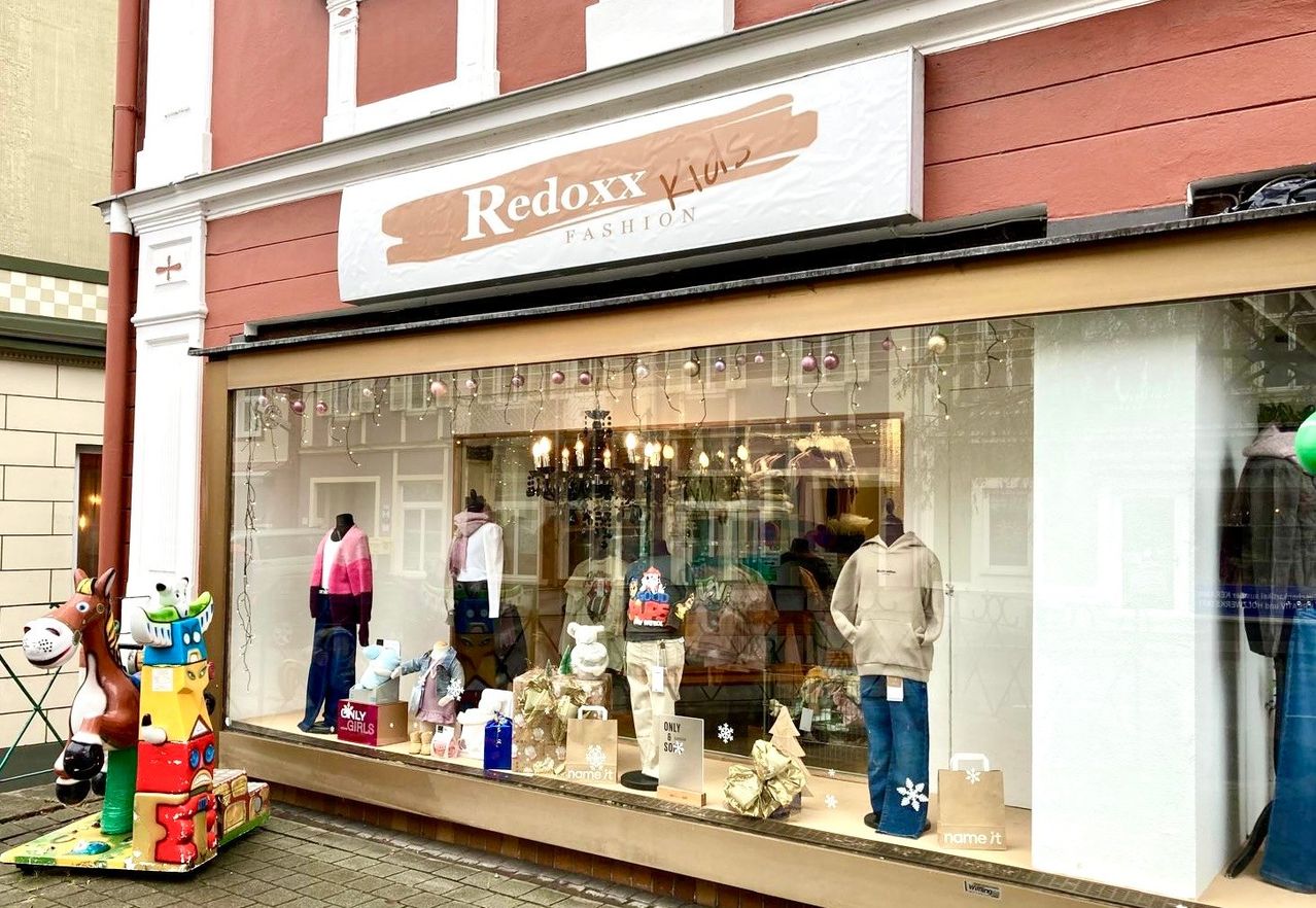 Ein Schaufenster mit einem Schild, das Redoxx Kids Fashion anzeigt, zeigt Winterkleidung. Mannequins präsentieren Jacken, Hoodies und Jeans. Das Fenster ist mit festlichen Dekorationen geschmückt.