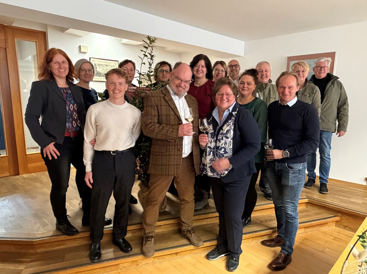 Eine Gruppe von Menschen versammelt sich in einem Raum und hält Weingläser in den Händen. Sie stehen auf einem Holzboden und lächeln für ein Foto. Sie tragen Winterkleidung. Ein Topf steht links. Der Raum hat weiße Wände und Decke.