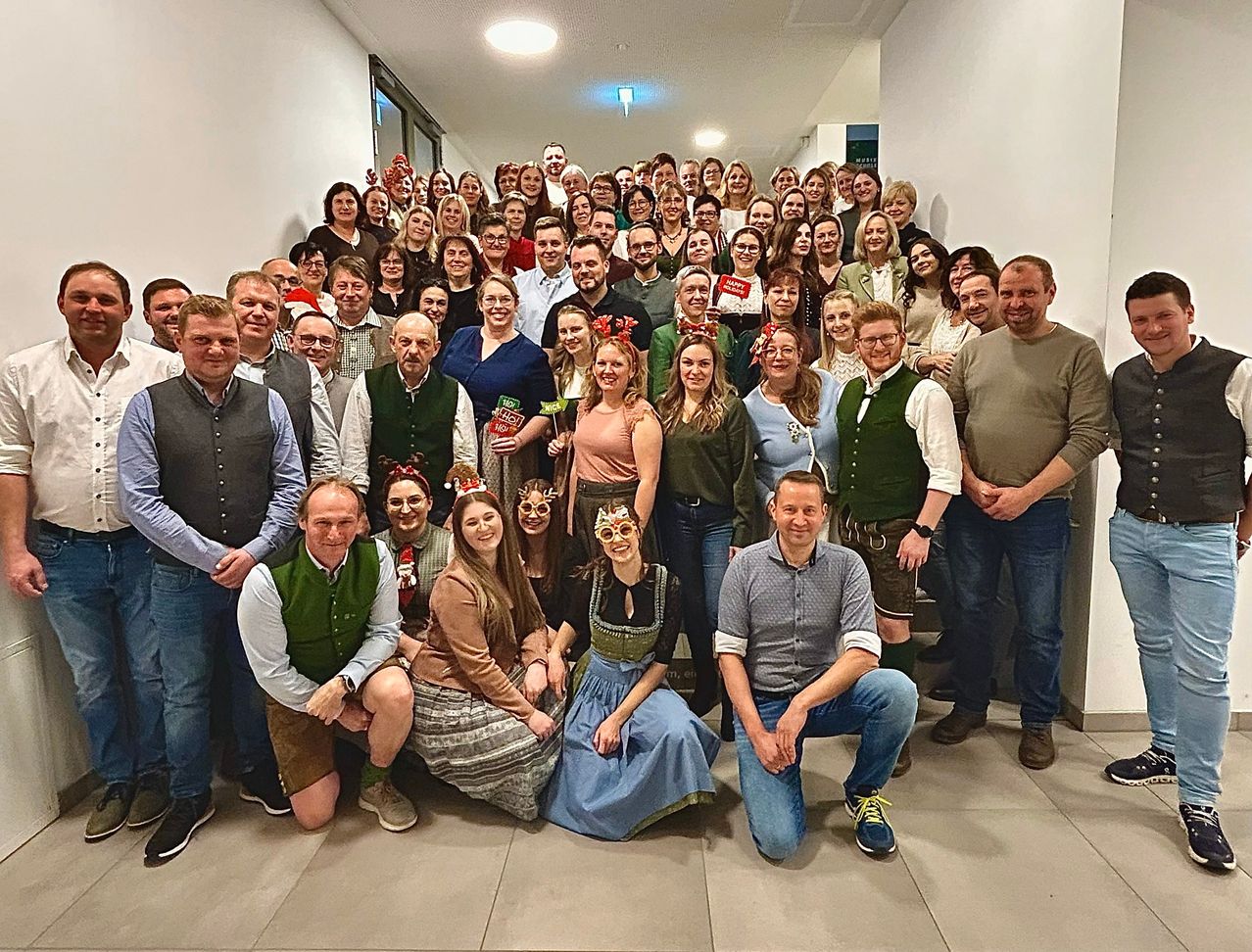 Eine Gruppe von Menschen in festlicher Kleidung ist für ein Foto versammelt. Sie stehen und sitzen in einem Flur, einige tragen Brillen und Mützen. Sie alle lächeln für die Kamera.