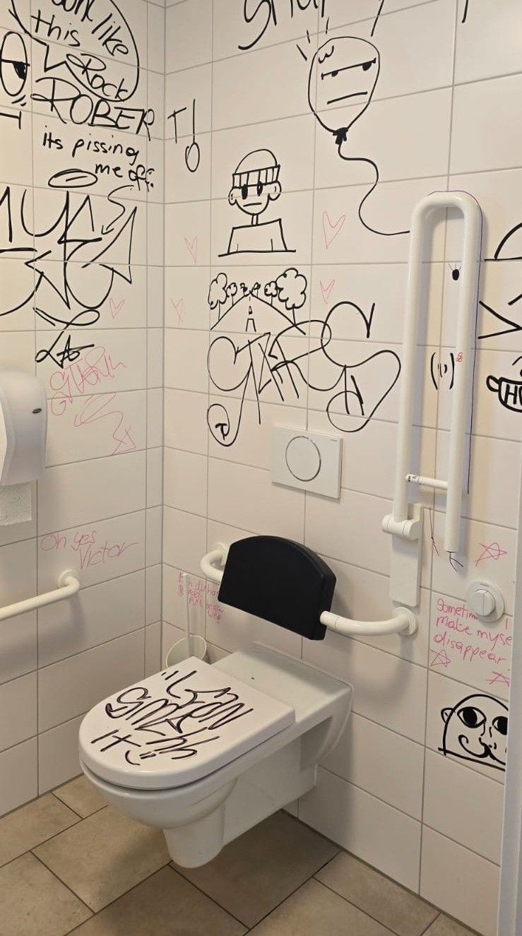 Ein Badezimmer mit weißen Fliesen und Graffiti. Eine Toilette mit Graffiti auf dem Sitz und ein weißer Toilettenpapierhalter. Die Wände haben Graffiti, einschließlich eines rosa Herzens und des Wortes 'Smell'. Ein weißer Haltegriff ist an der Wand montiert.